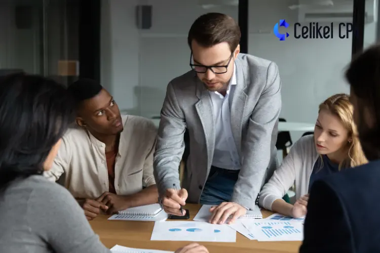 A business meeting at Celikel CPA showcasing a young and skilled workforce. A man in glasses and a gray blazer is explaining a chart to his colleagues. Three team members are attentively listening and reviewing documents with financial graphs. The "Celikel CPA" logo is visible in the background.